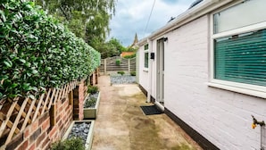 Un étroit sentier mène à un bâtiment blanc orné d’un volet de fenêtre vert, le tout étant entouré d’un mur de briques et d’une haie.