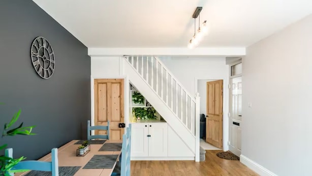 Un coin repas avec une table et des chaises en bois, une horloge au mur et un escalier menant à une autre pièce.