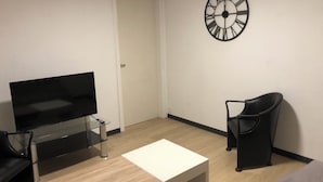 A modern living room with a flat-screen TV on a glass table, a white coffee table, a black leather chair, and a large wall clock. - Gîte 'A deux pas du ciné', located in the center of Maîche (Maîche)