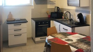 A compact kitchen with white cabinets, a black countertop, a stove, a microwave, and a coffee maker. There is a window with blinds, a small table with a red tablecloth, and a chair. - Gîte 'A deux pas du ciné', located in the center of Maîche (Maîche)
