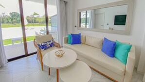 A modern living room with a sofa, two round tables, a wicker chair, and a large window offering a view of a pool and greenery.