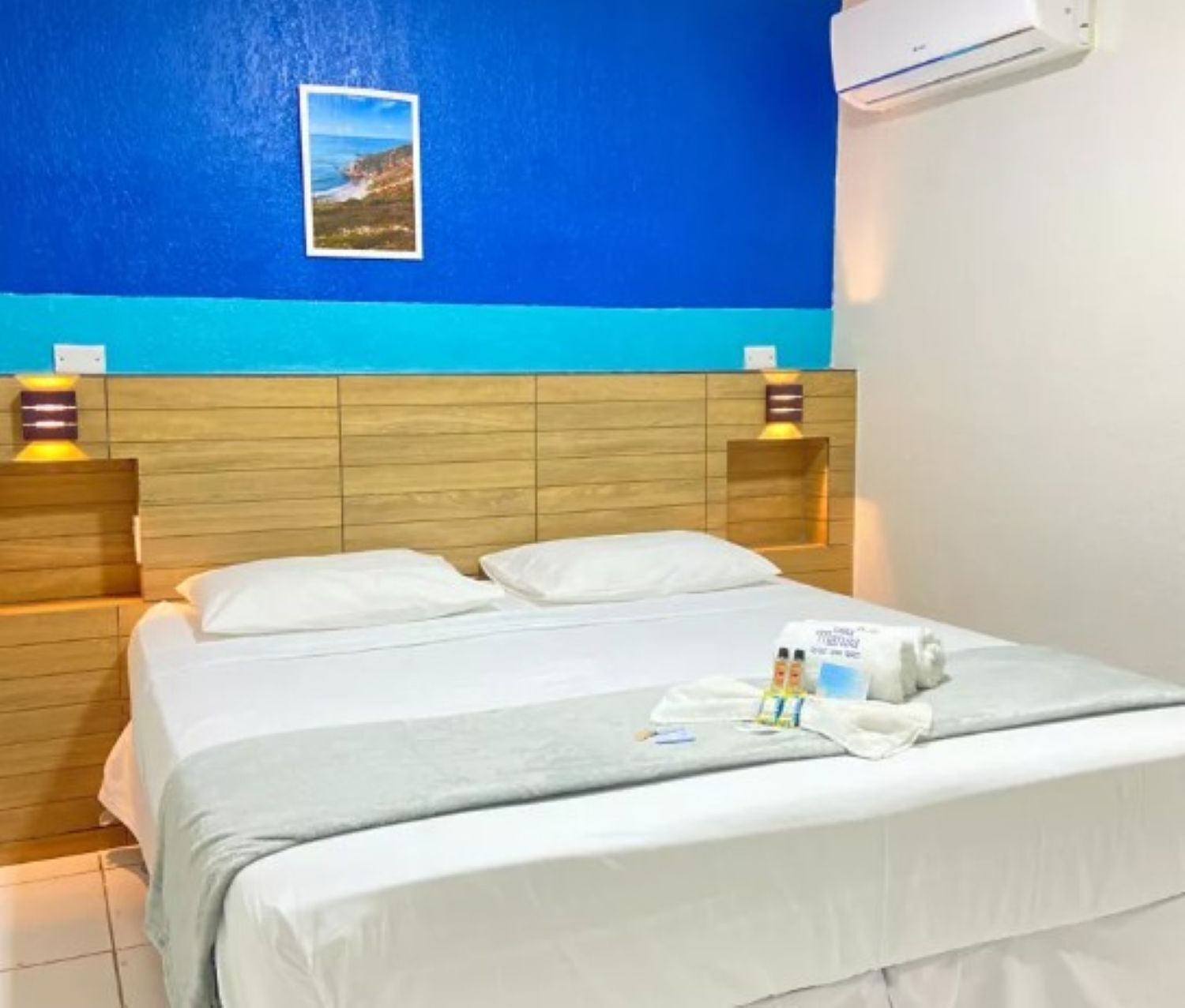 Habitación de hotel con cabecera de madera, cama con sábanas blancas, aire acondicionado de pared y un cuadro enmarcado sobre la cama.