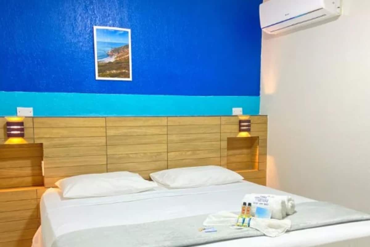 Une chambre d’hôtel avec une tête de lit en bois, un lit recouvert de draps blancs, un climatiseur fixé au mur et un tableau encadré au-dessus du lit.