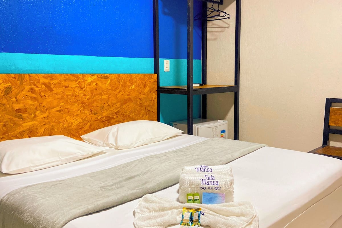 Une chambre d’hôtel avec un lit, du linge de lit blanc, un mur bleu et orange, une tête de lit en bois, une petite table et un mini-bar.
