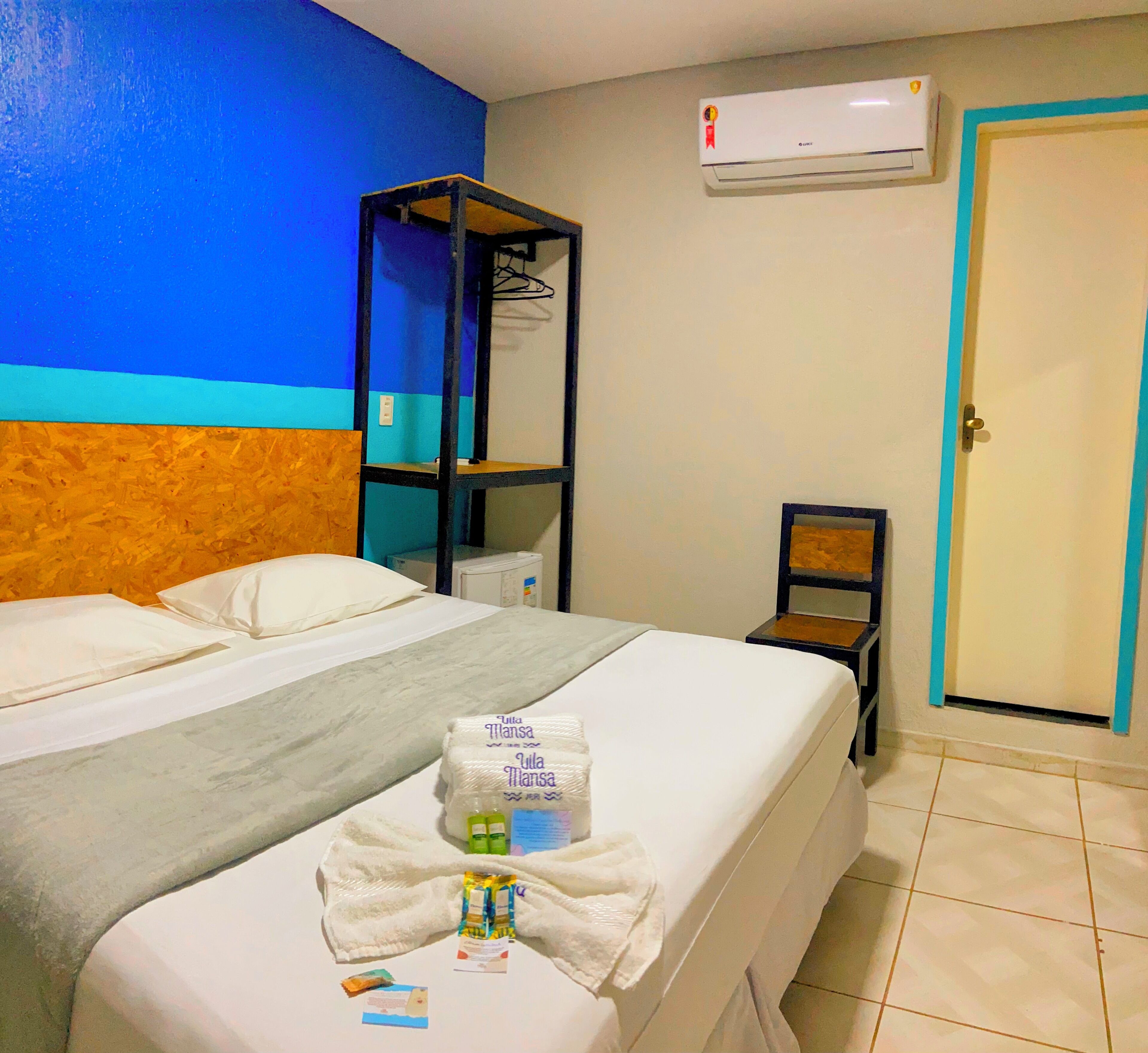 A hotel room with a bed, a small table with toiletries, a shelf, and a door.