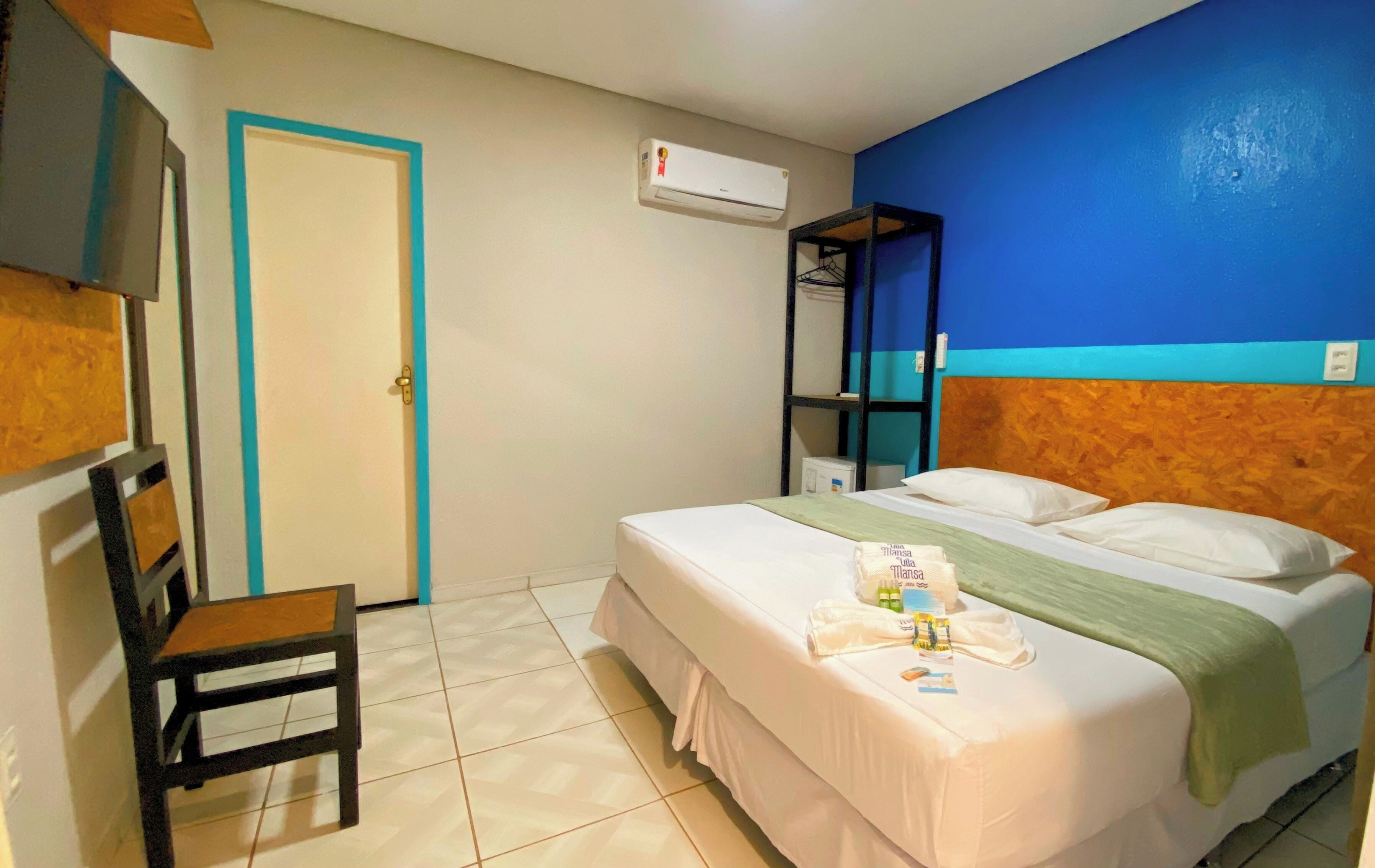 A hotel room with a bed, a chair, a shelf, and an air conditioning unit.
