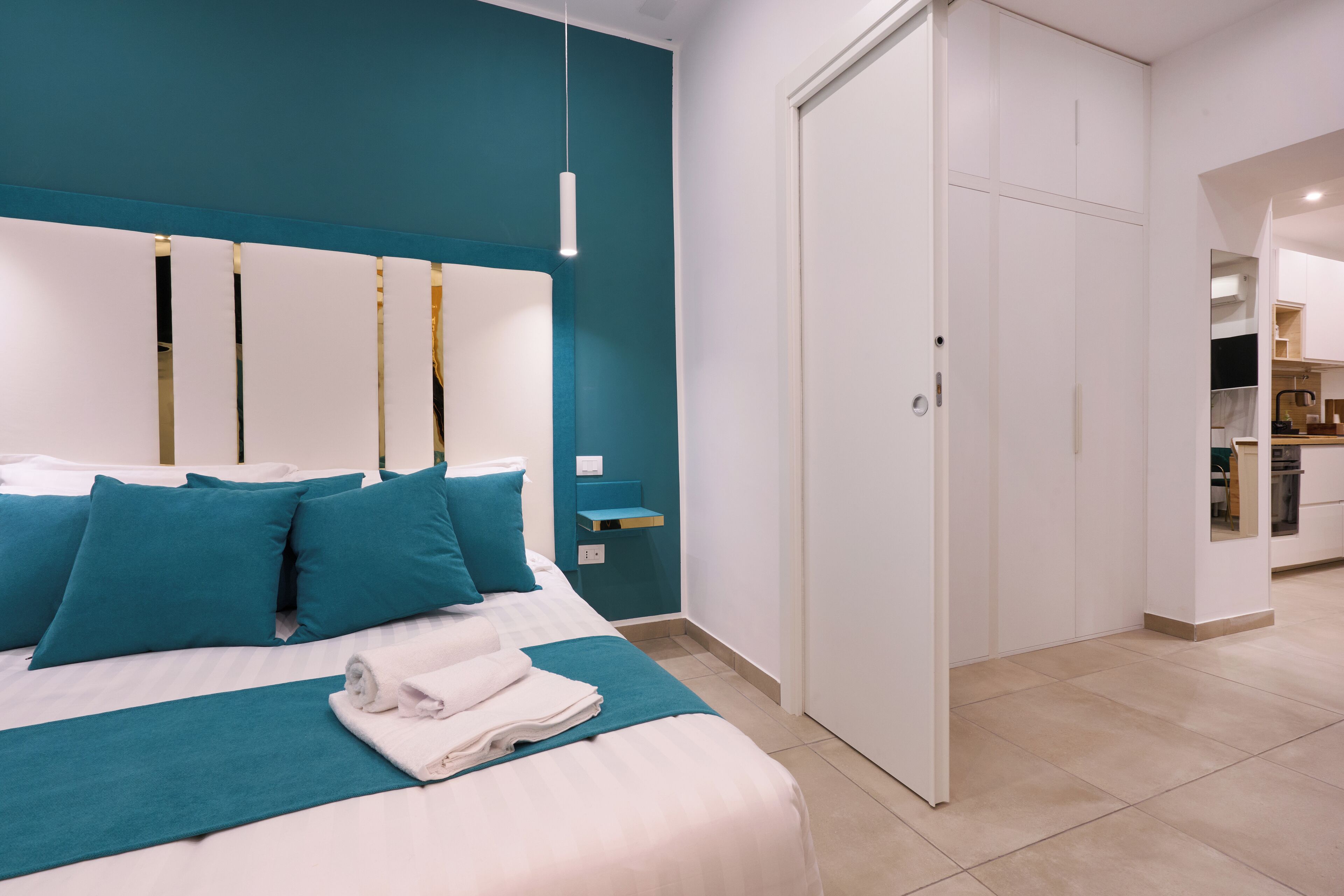 A modern bedroom with a teal accent wall, a large bed with blue bedding, and a white wardrobe.