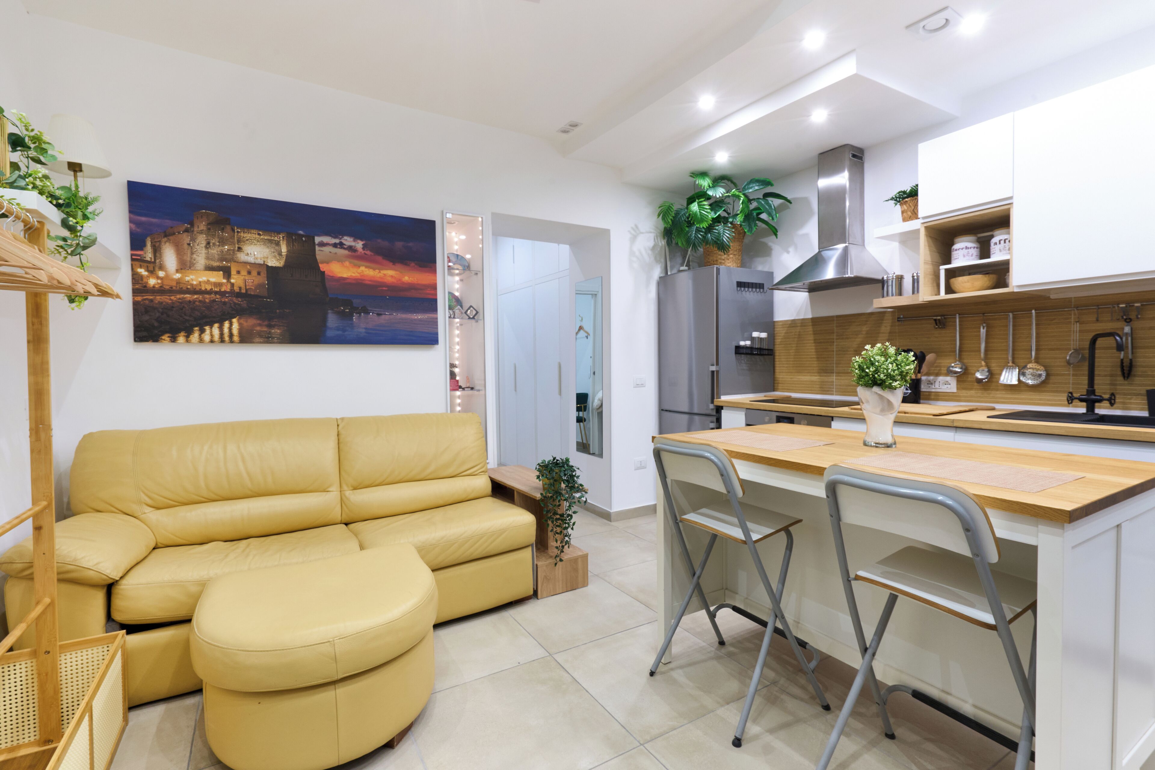 A modern kitchen with a built-in island, white cabinetry, and stainless steel appliances. There is a yellow leather sofa and a small dining area with two chairs.