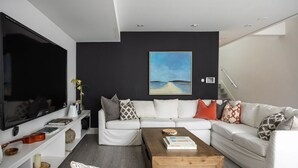 Ein modernes Wohnzimmer mit großem Flachbildfernseher, einer weißen Sofagarnitur, einem Couchtisch und einem Bild an der Wand.