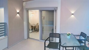 A modern dining area with a glass table and chairs, a sliding glass door leading to a dining room, and two wall-mounted lights.