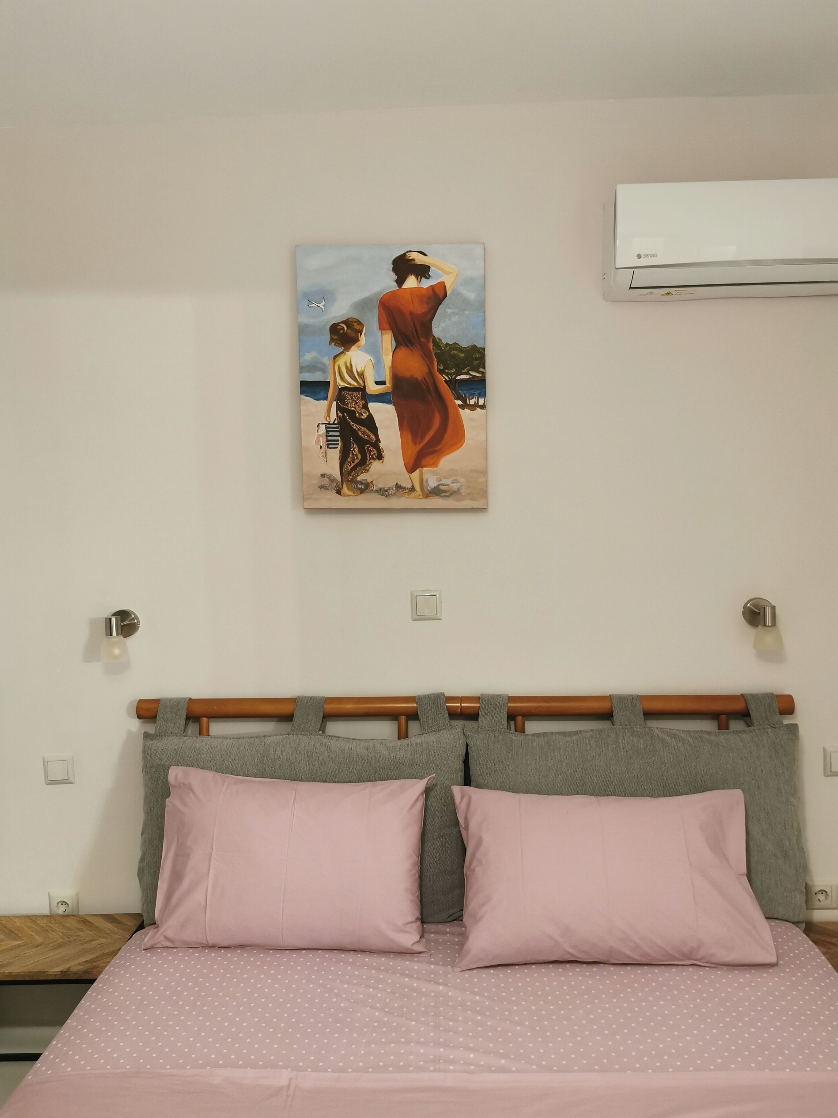 Een slaapkamer met een bed, twee kussens, een hoofdbord, een wandairconditioner en een schilderij van een vrouw en kind op het strand.