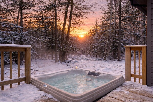 Un bain à remous dans une forêt enneigée, avec un coucher de soleil en arrière-plan.