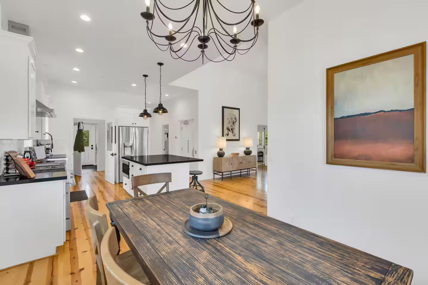 A modern kitchen with a wooden dining table, a chandelier, and a painting on the wall.