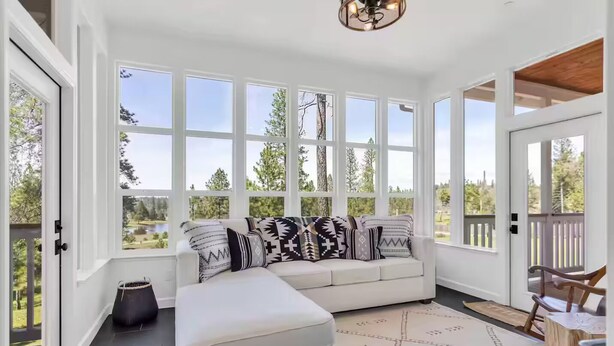 A sunlit room with a white sofa, patterned cushions, and a view of a lake through large windows.