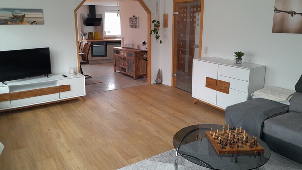 Una sala de estar moderna con un televisor de pantalla plana, una mesa de centro de vidrio con un juego de ajedrez, un sofá gris y una cocina al fondo.
