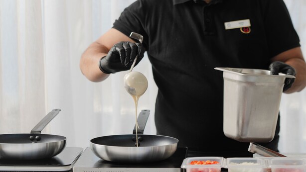 A person in a black uniform pouring a liquid from a ladle into a pan on a stove. - Serenity Suite Caldera View with Hot Tub (Thira - Santorini)
