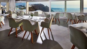 A modern dining area with white tables and chairs, large windows offering a view of a coastal landscape. - Serenity Suite Caldera View with Hot Tub (Thira - Santorini)