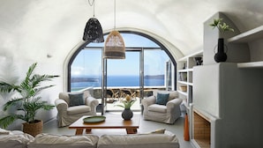 A spacious living area with a large arched window offering a view of the sea, a cozy seating arrangement, and a fireplace. - Grand Honeymoon Villa with Sea View and Hot tub (Thira - Santorini)