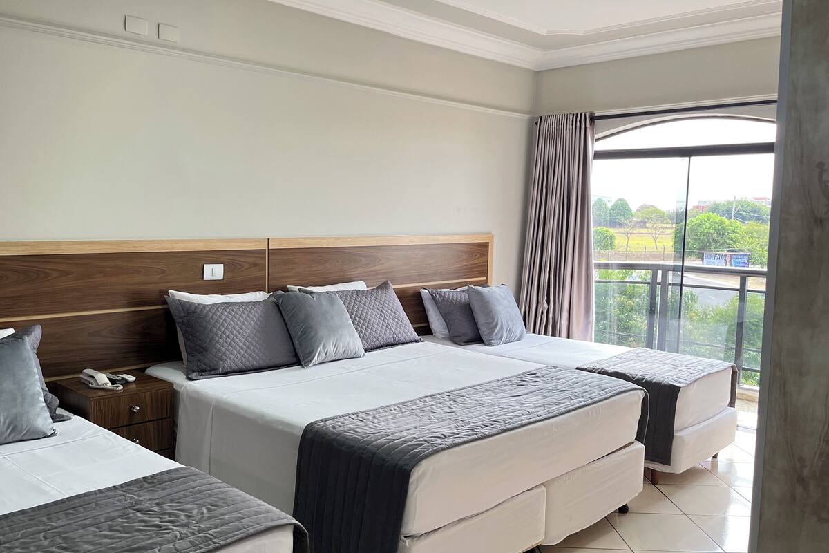 A hotel room with two beds, a wooden headboard, a nightstand, and a view of greenery through the window.