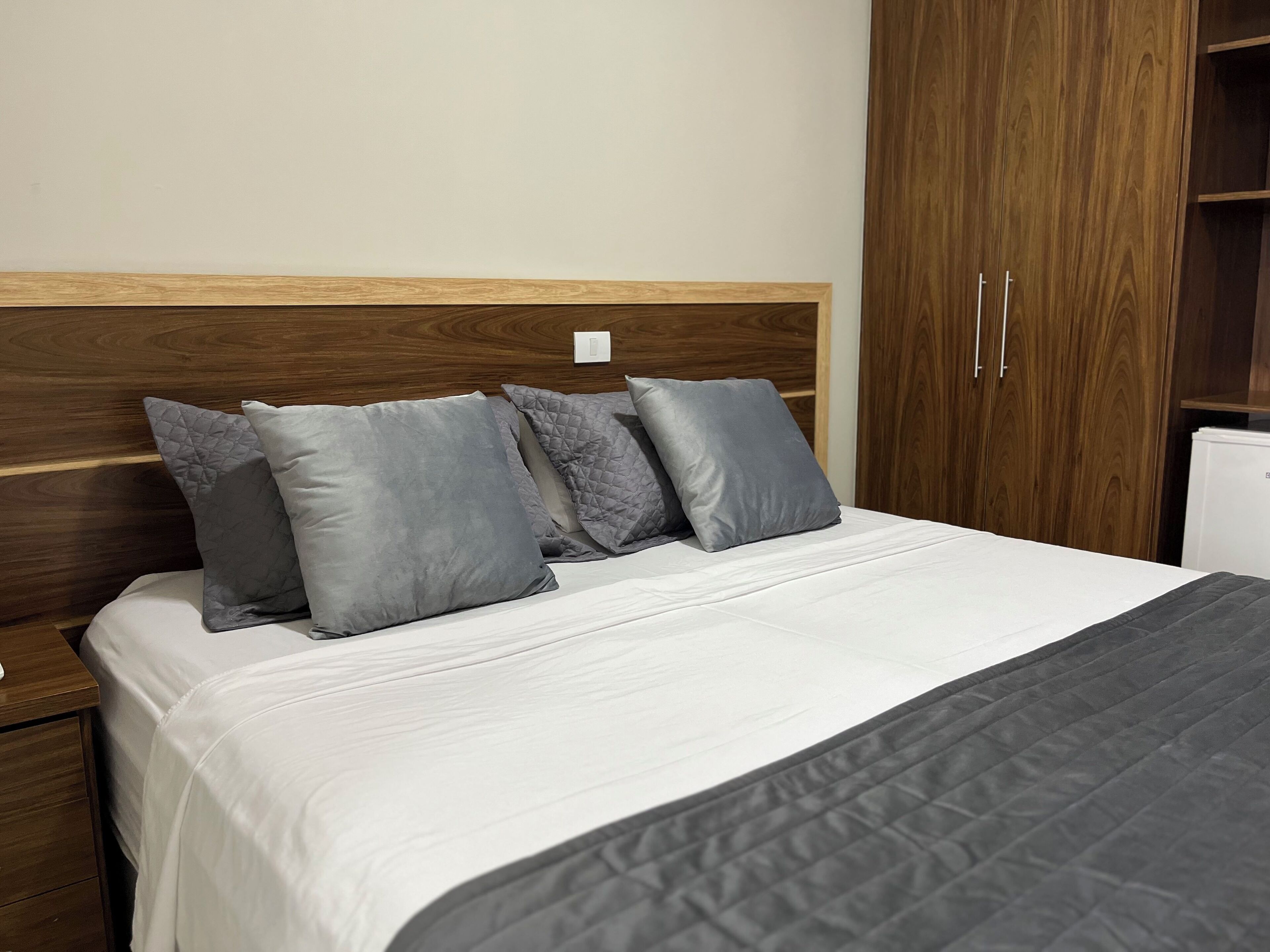 A neatly made bed with a white duvet and grey quilted pillows, a wooden headboard, and a wooden wardrobe with two doors.