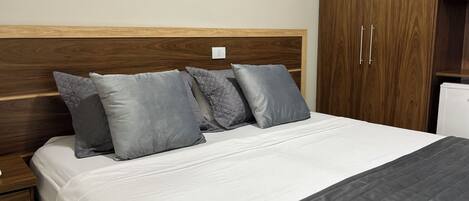 Uma cama bem arrumada com um edredom branco e almofadas cinza acolchoadas, uma cabeceira de madeira e um guarda-roupa de madeira com duas portas.