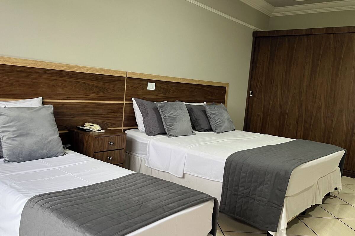 A hotel room with two beds, each with white bedding and grey pillows. There is a wooden headboard, a nightstand with a telephone, and a wardrobe.
