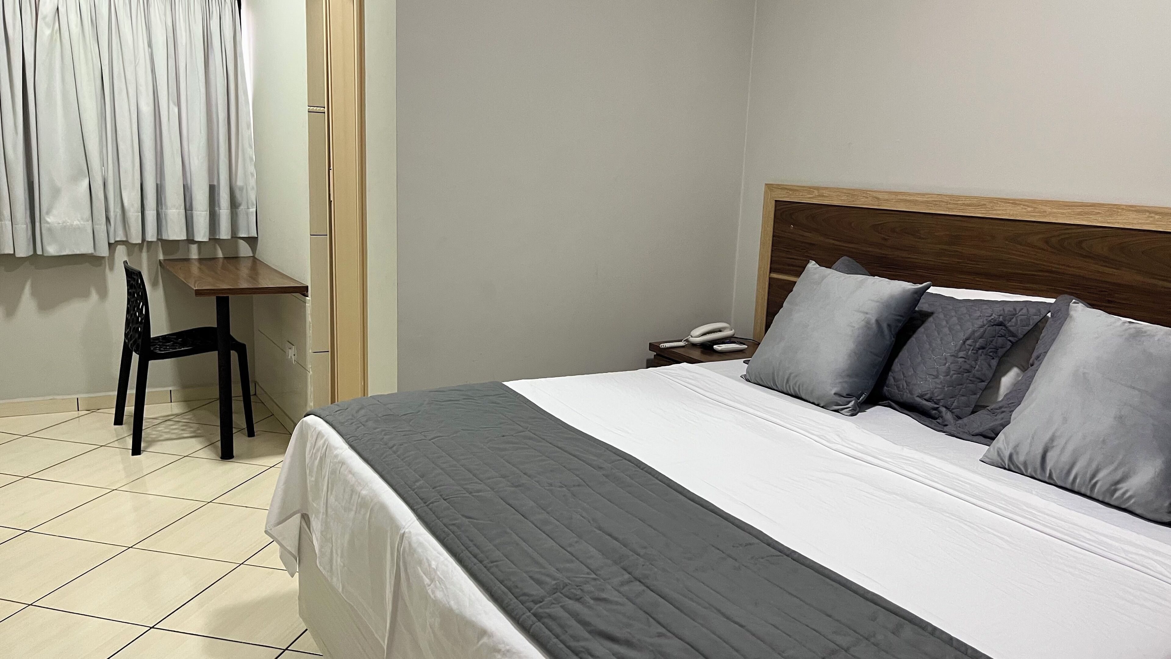 A hotel room with a bed, a desk, a chair, and a nightstand with a phone.