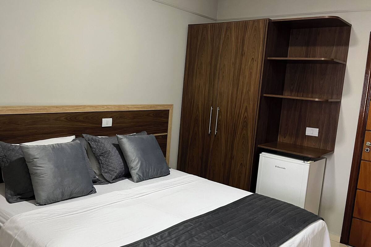 A bedroom with a bed, grey pillows, a wooden headboard, a wardrobe, and a small white appliance.