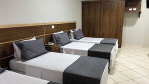 A hotel room with three beds, each with a white and black bedspread, and grey pillows. There is a wooden headboard, a wardrobe, and a small shelf with items on it. - Gutierrez Hotel (Americana)
