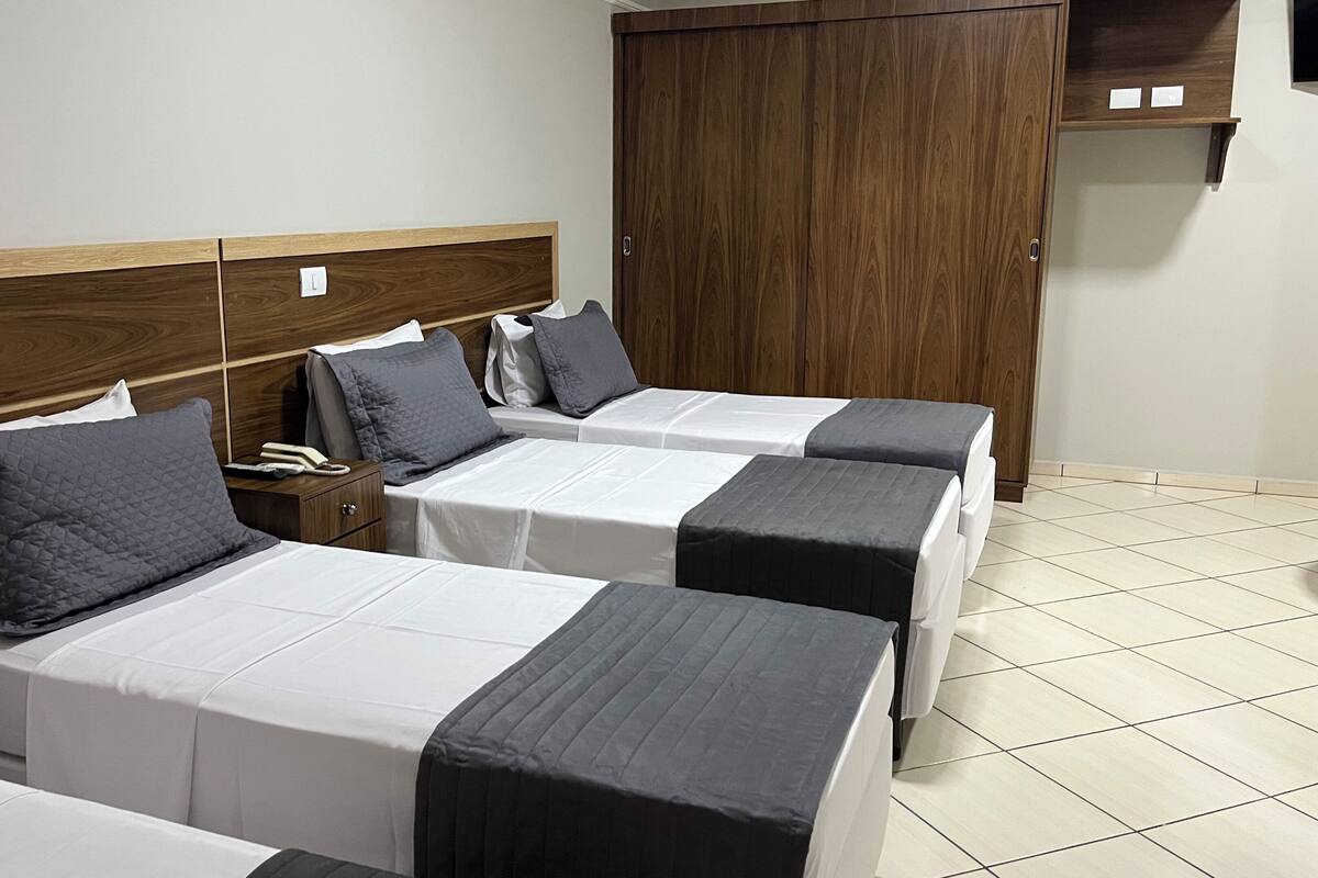 A hotel room with three beds, each with a white and black bedspread, and grey pillows. There is a wooden headboard, a wardrobe, and a small shelf with items on it.