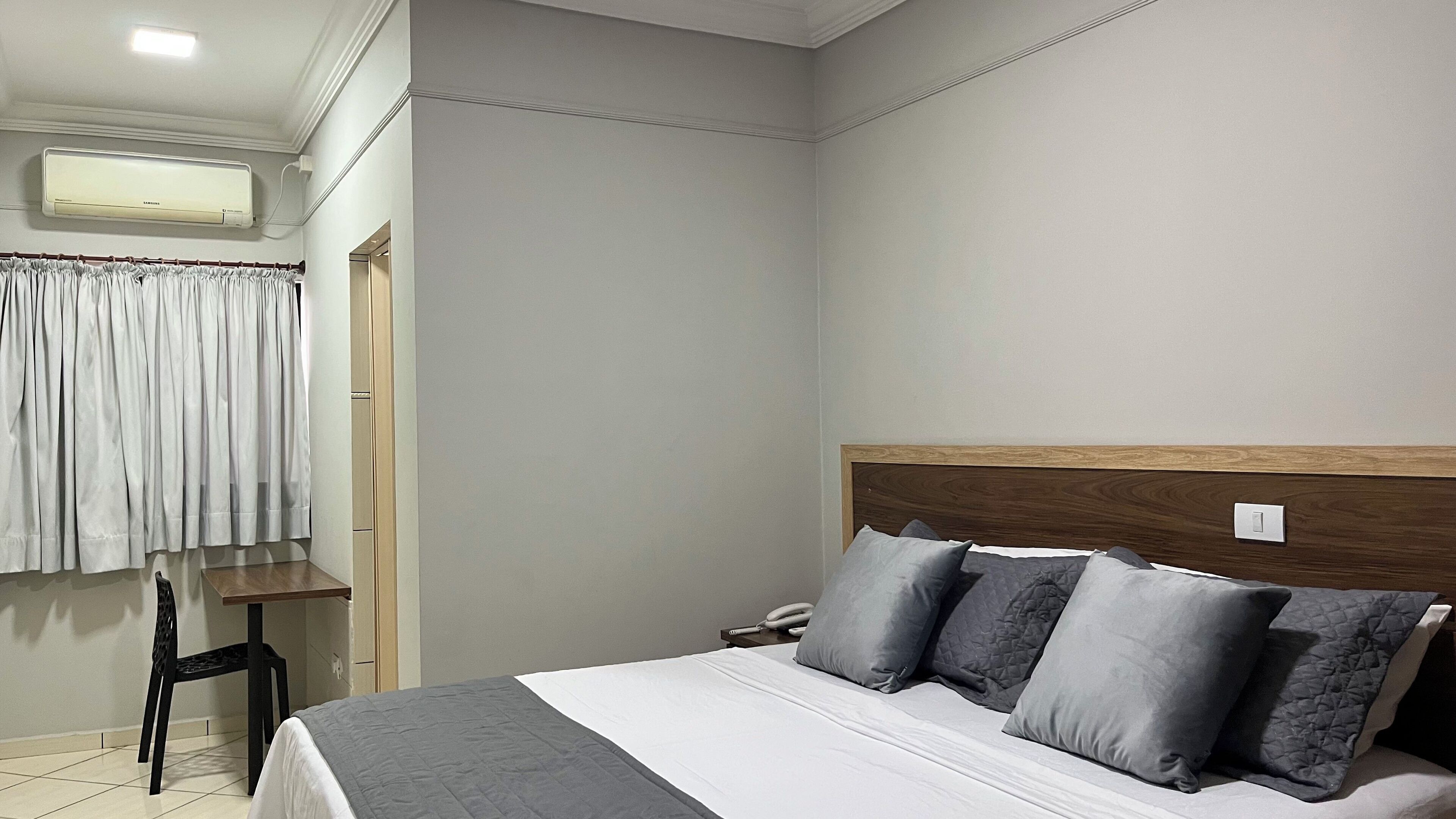 A hotel room with a bed, bedside table, chair, and an air conditioning unit.
