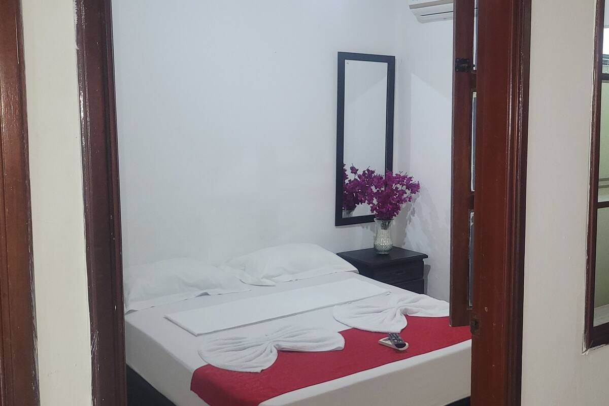 A neatly made bed with white linens and a red blanket, a small bedside table with a vase of purple flowers, and a mirror on the wall.