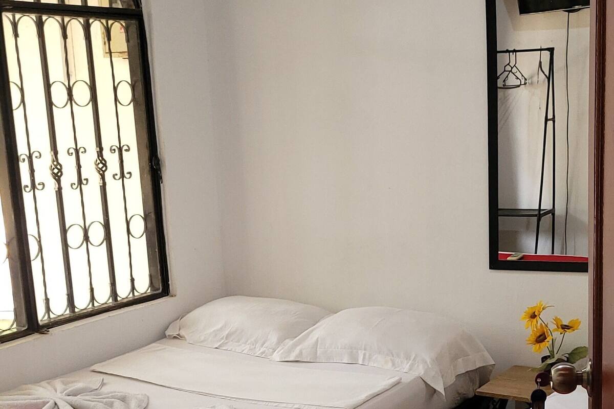 A room with two beds, each with a red bedspread and white linens. There is a window with bars, an air conditioning unit on the wall, and a mirror with a black frame.