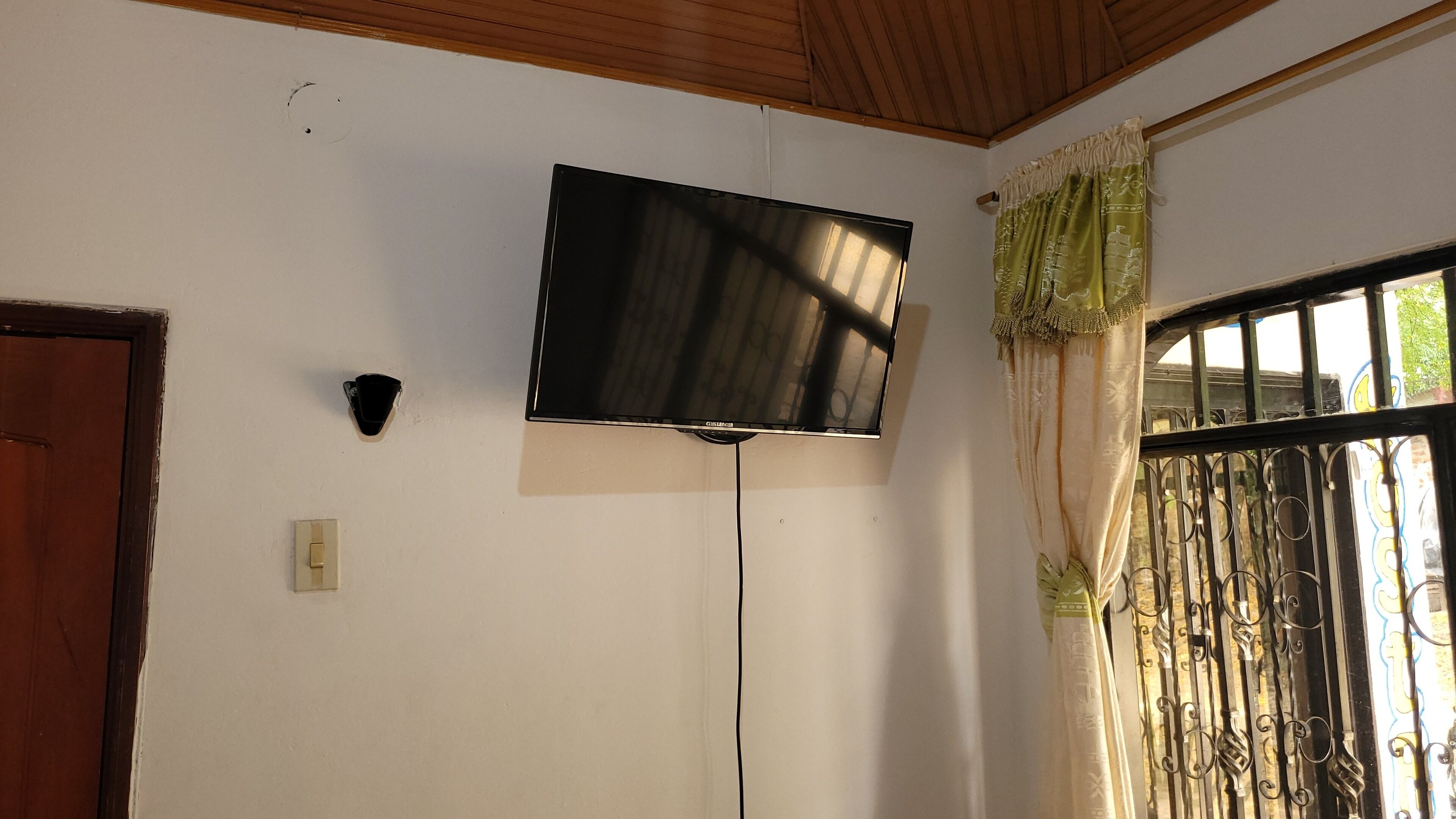 A room with a mounted television on the wall, a door, and a window with decorative curtains.