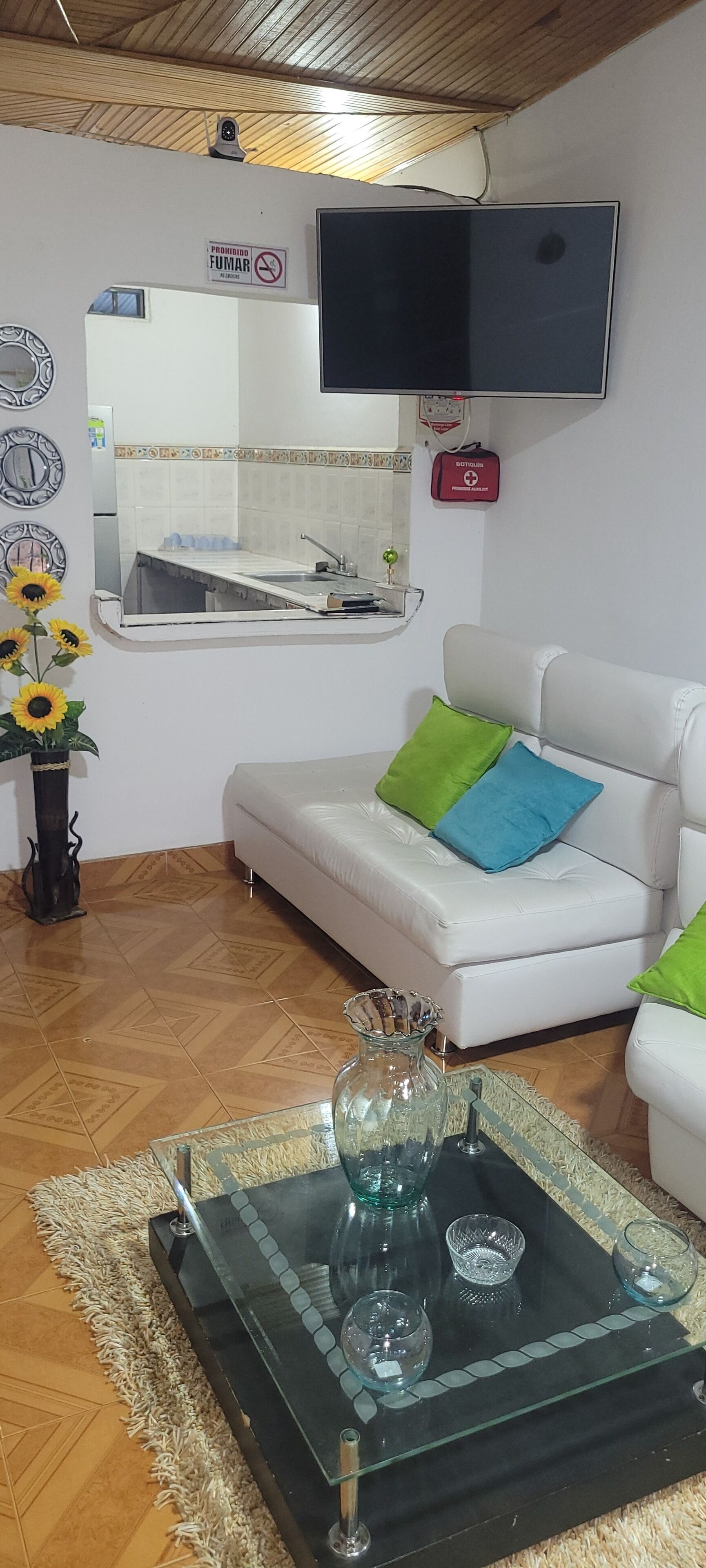 Ein Wohnzimmer mit einem weißen Sofa, einem Glastisch, einem an der Wand befestigten Flachbildfernseher und einer Küchenzeile mit Spüle und Fliesenrückwand.