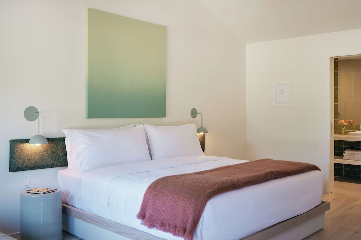 A modern bedroom with a large bed, two bedside lamps, a green abstract painting, and a wooden floor.