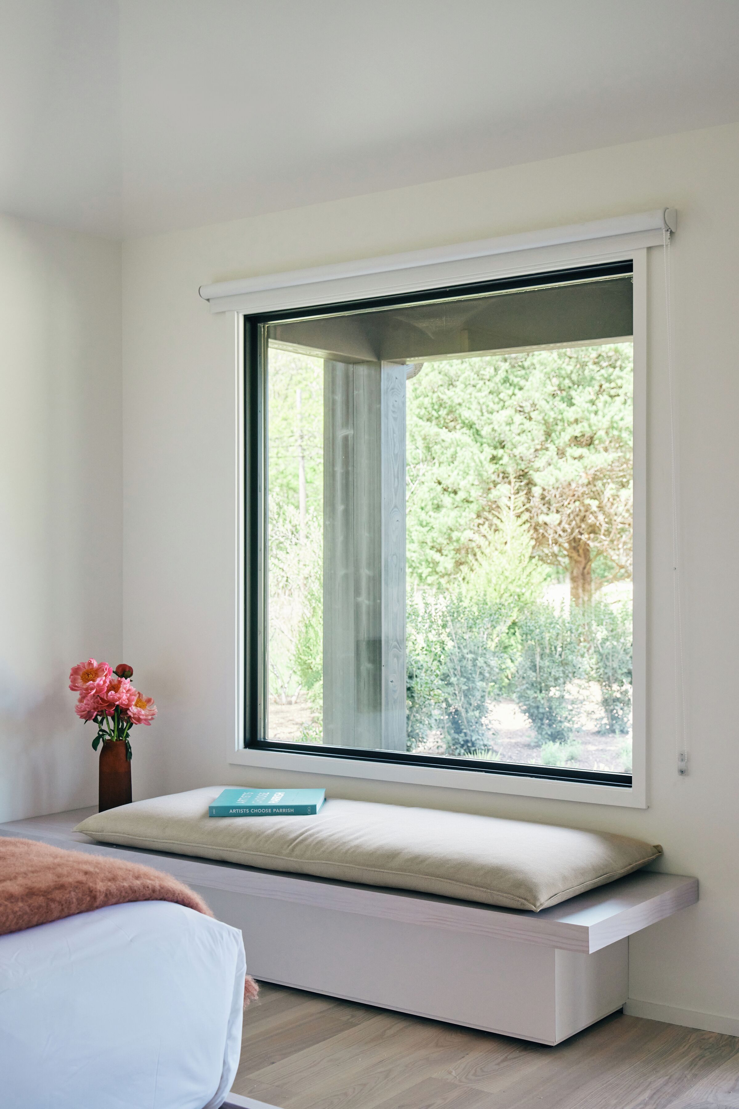 A room with a built-in bench under a large window, a bedside table with a book, and a vase with flowers.