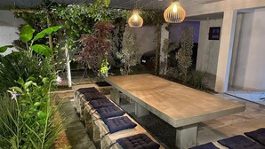 Een buitenterras met een lange tafel en banken, omgeven door weelderige begroeiing en hangende lampen.