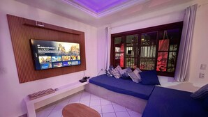 Een woonkamer met een aan de muur bevestigde televisie, een bank, een klein houten tafeltje en een raam met uitzicht op beplanting.