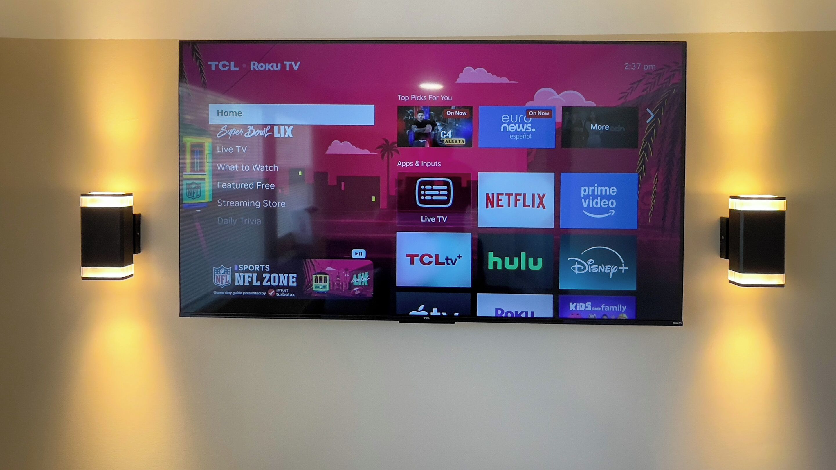 A TCL Roku TV mounted on a wall with a menu interface displaying various streaming services and options.
