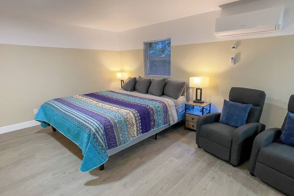 Ein Schlafzimmer mit Bett, zwei Stühlen, einem Nachttisch, einer Lampe und einer Klimaanlage.