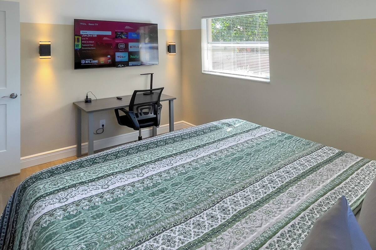 Ein Schlafzimmer mit Bett, Schreibtisch, Stuhl, einem an der Wand befestigten Fernseher und einem Fenster mit Jalousien.