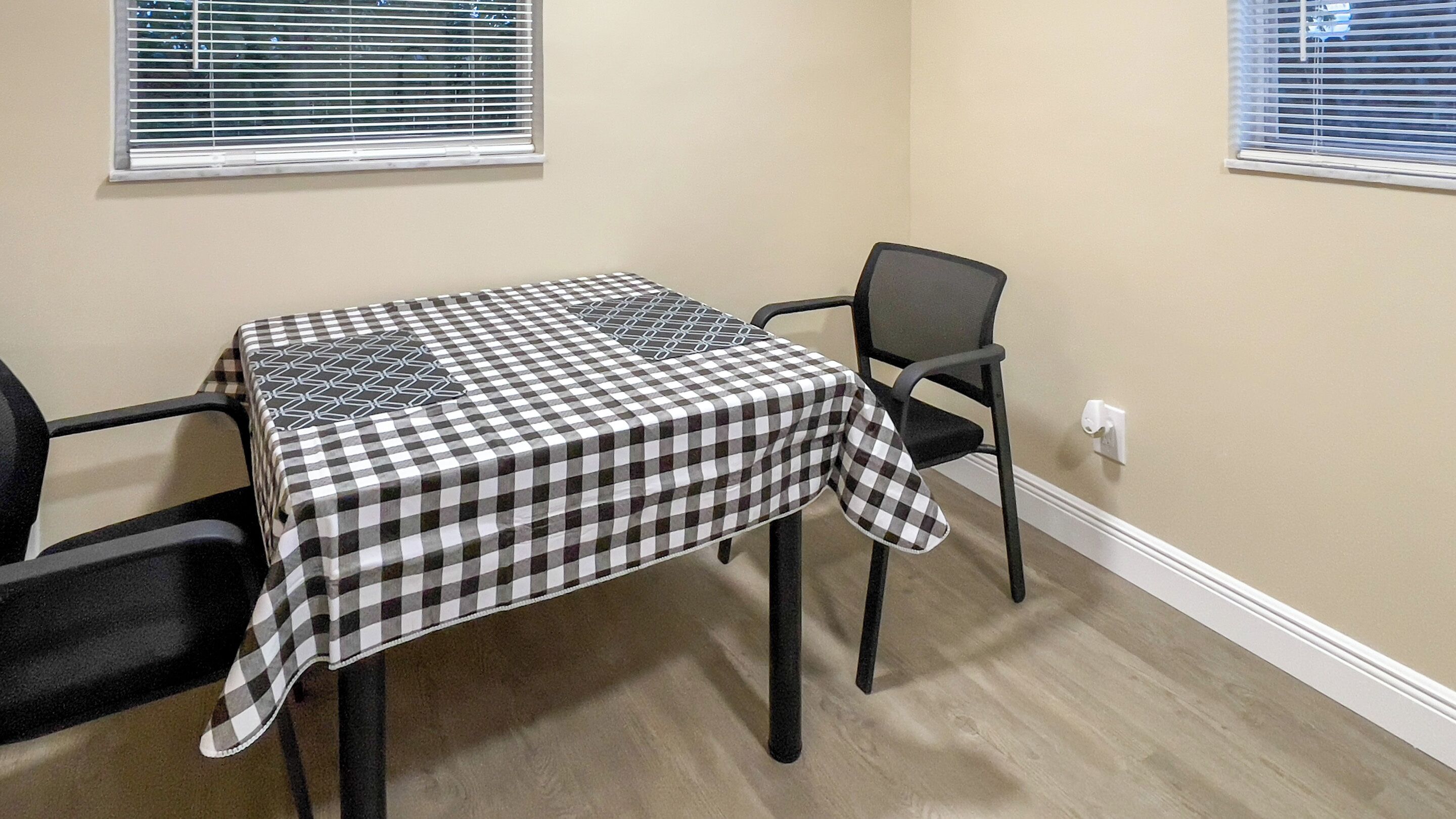A dining area with a checkered tablecloth, black chairs, and a window with blinds.