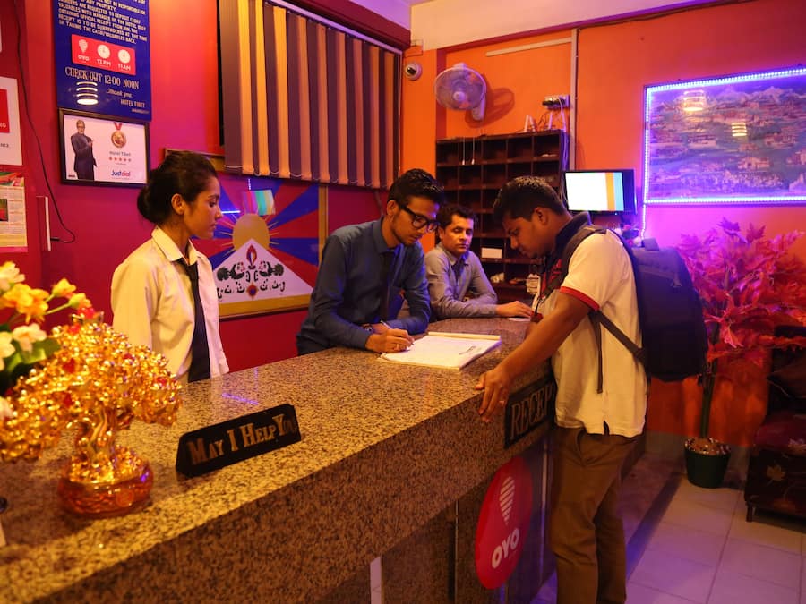 A hotel reception desk with a sign reading "May I Help You?" and a "Reception" sign. There are posters on the wall, a fan, and a potted plant.