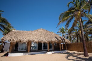 Una estructura de techo de paja con amplios ventanales, rodeada de palmeras y una playa arenosa.