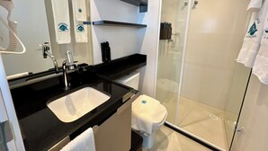 A modern bathroom with a black countertop, white sink, and a glass-enclosed shower. There are towels hanging on the wall and a toilet with a towel on it. - RN STUDIO - APTO 905 (Maceió)