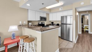A modern kitchen with white cabinets, a stainless steel refrigerator, a microwave, and a coffee maker. There is a small island with stools and a table with a lamp. - Oceanfront Oasis: Your Ideal 1BR Condo Escape (Myrtle Beach)