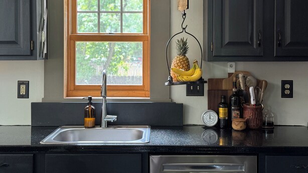 A kitchen with a window, a hanging fruit basket, a sink, and various kitchen items on the counter. - THE MILL HOUSE - Charming Historic Cottage 1.5 miles from the US Open (Oakmont)