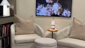 A modern living room with a beige sofa, a small ottoman, a table with a lamp, and a bookshelf filled with books. - THE MILL HOUSE - Charming Historic Cottage 1.5 miles from the US Open (Oakmont)