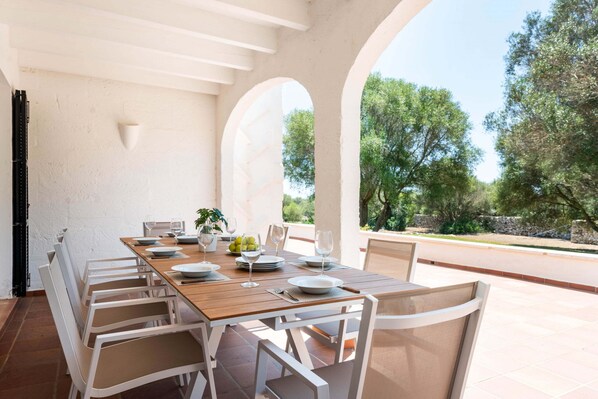 An outdoor dining area with a wooden table set for six, surrounded by white chairs, under a white ceiling with arches.